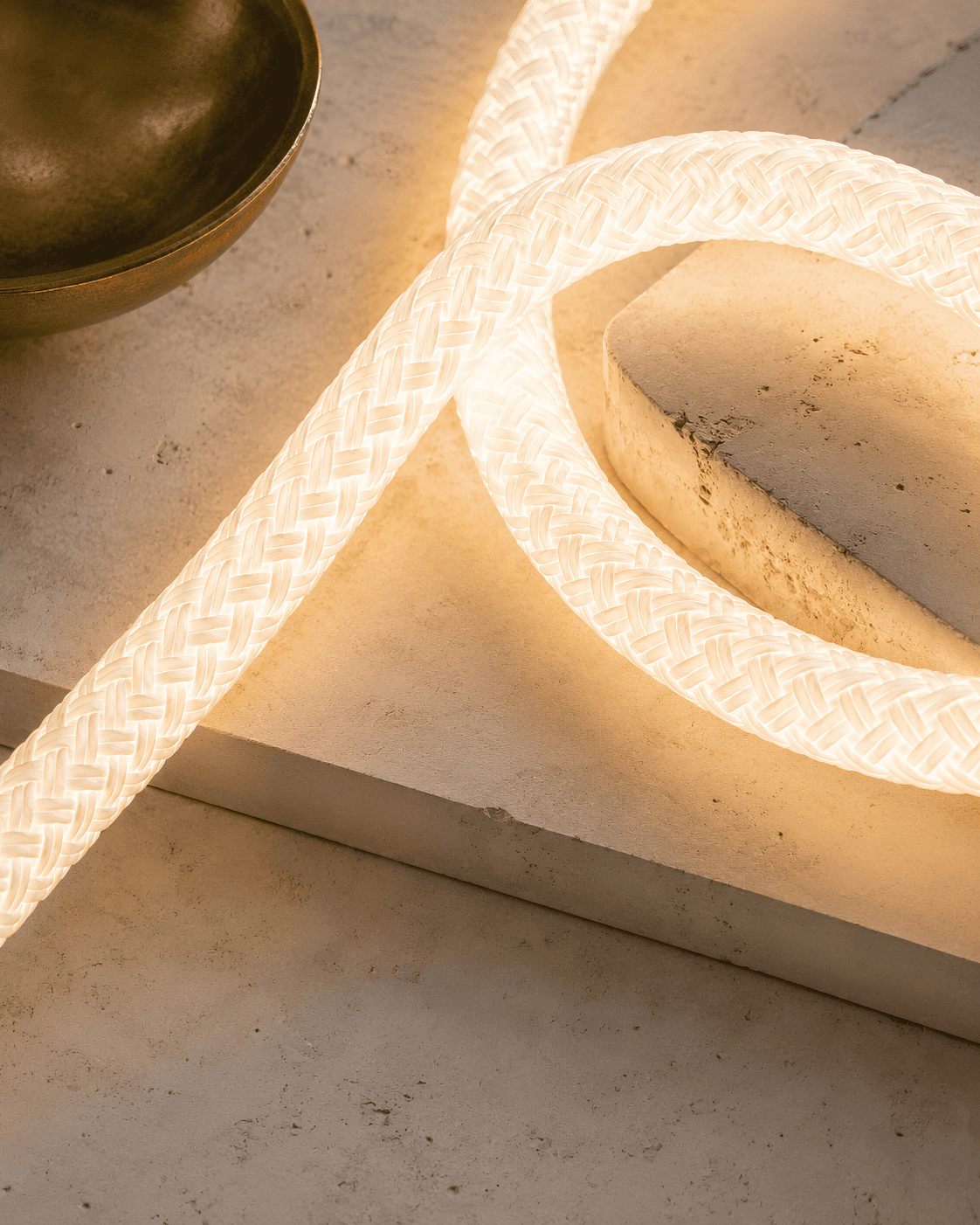 Illuminated braided cord looping across travertine stone beside a hand-finished brass bowl