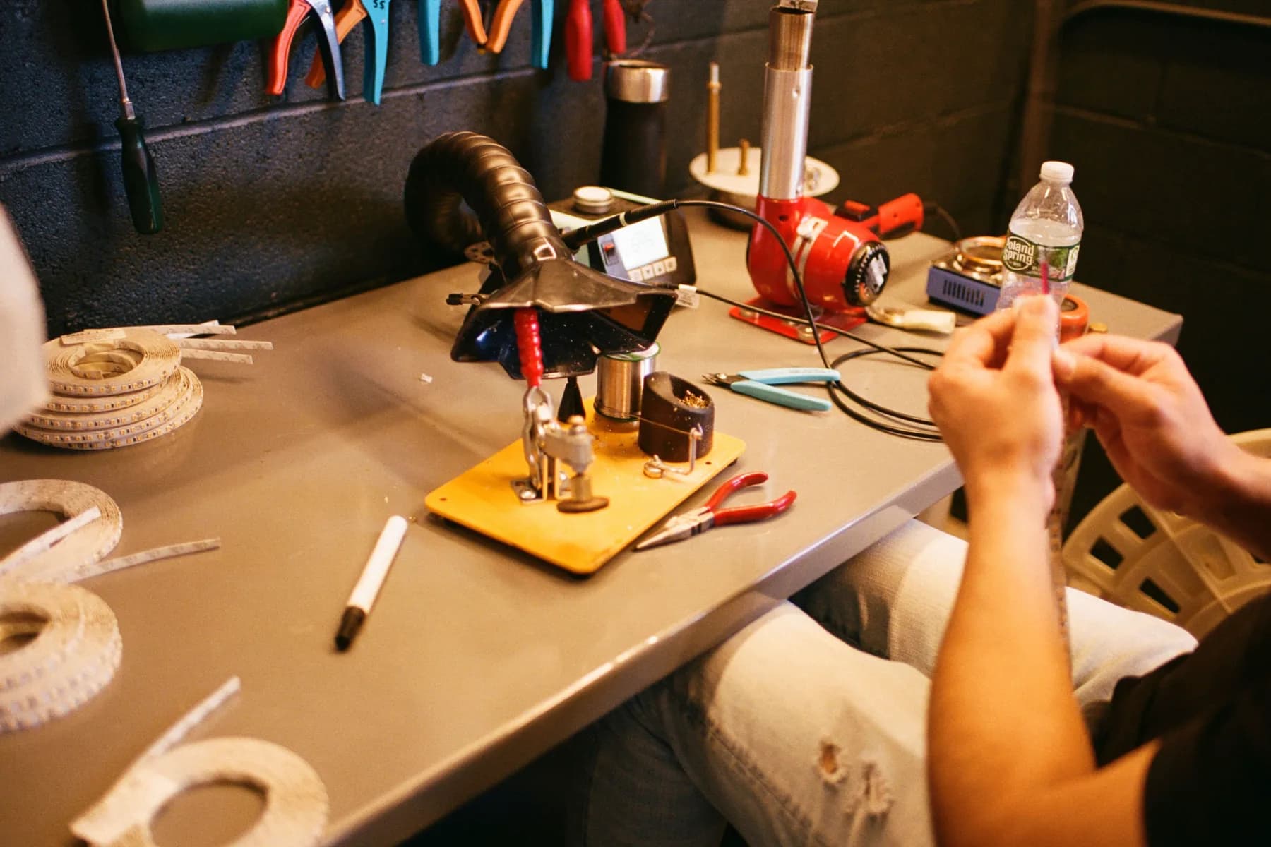 Hands working with wiring at a workbench with tools, LED reels, and a soldering station